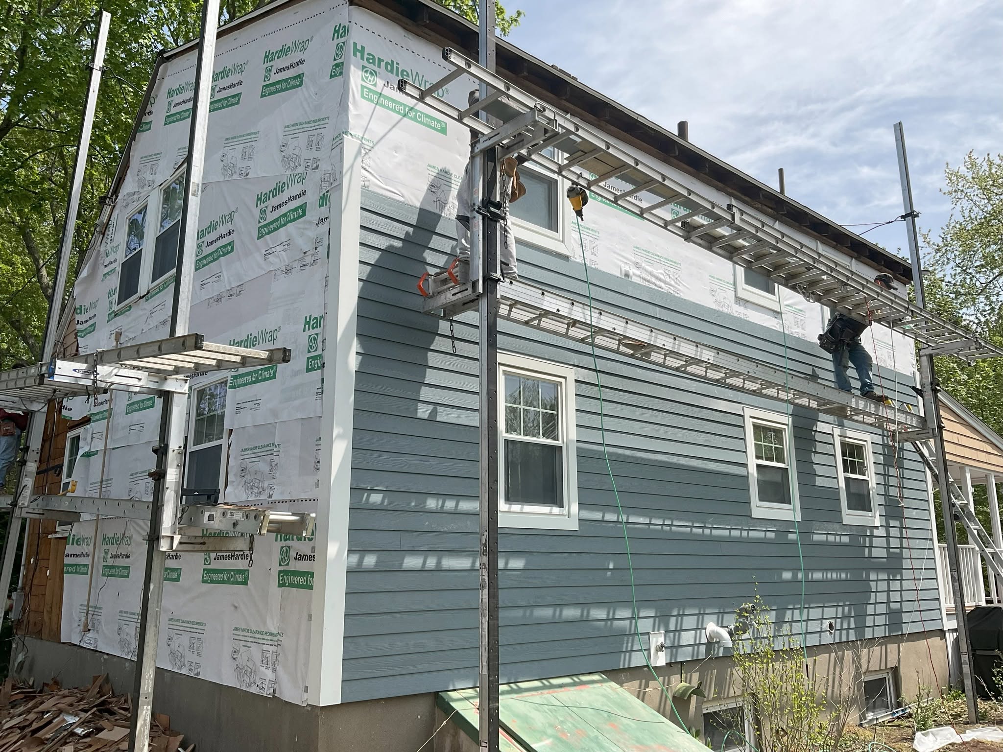 Construction workers on scaffolding installing blue-gray horizontal lap siding over house wrap on a two-story home.