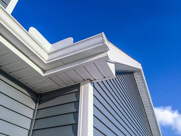 Low-angle close-up of white vinyl soffit, fascia, gutter, and corner trim on a house with gray siding against a bright blue sky.